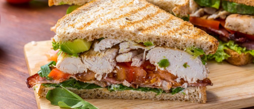 Broodje met kip, avocado en bacon op houten plank.