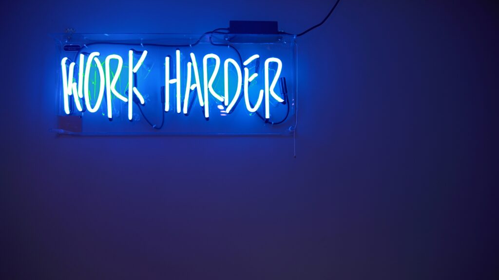 Blauw neonbord met de tekst 'Work Harder' op muur.