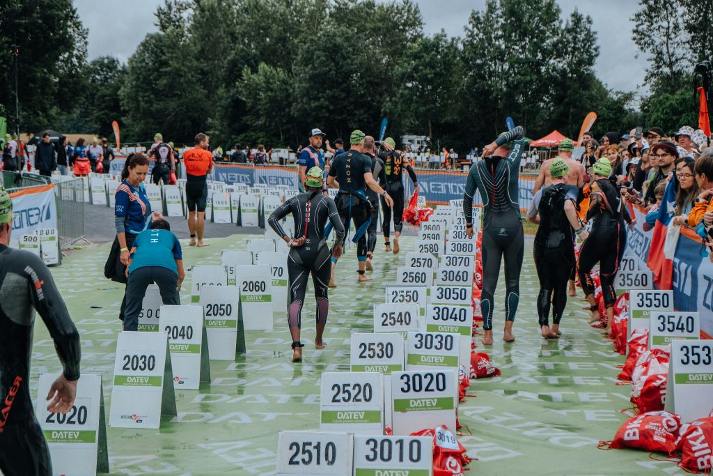 Triatleten in zwarte wetsuits staan tussen genummerde startborden op een groene mat voor een triatlon.