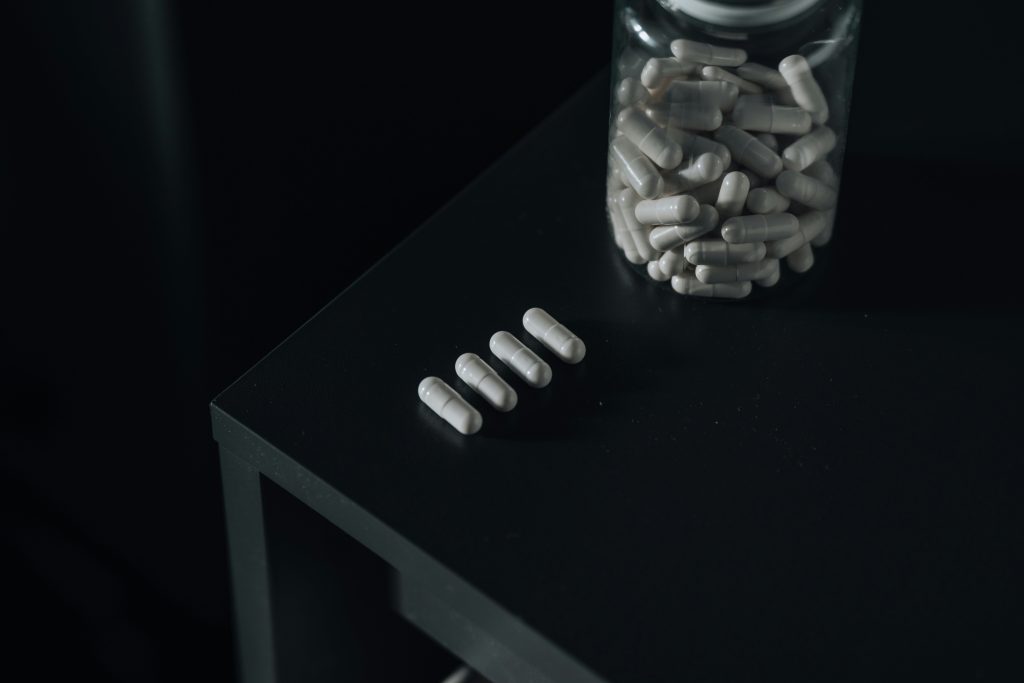 Witte capsules liggen naast een glazen pot op zwarte tafel.