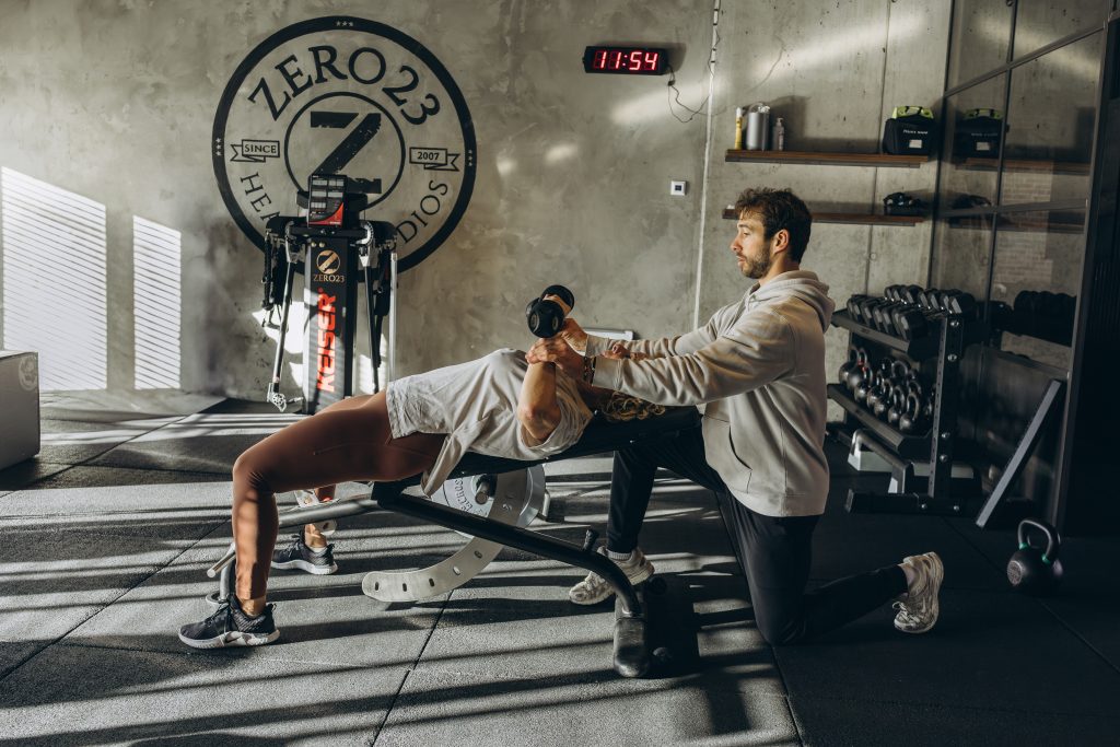 Vrouw doet bankdrukken met hulp van een trainer in sportschool.