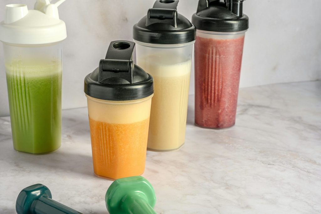 Vier drinkbekers met verschillende kleuren mixdranken en twee dumbbells op een marmeren oppervlak.