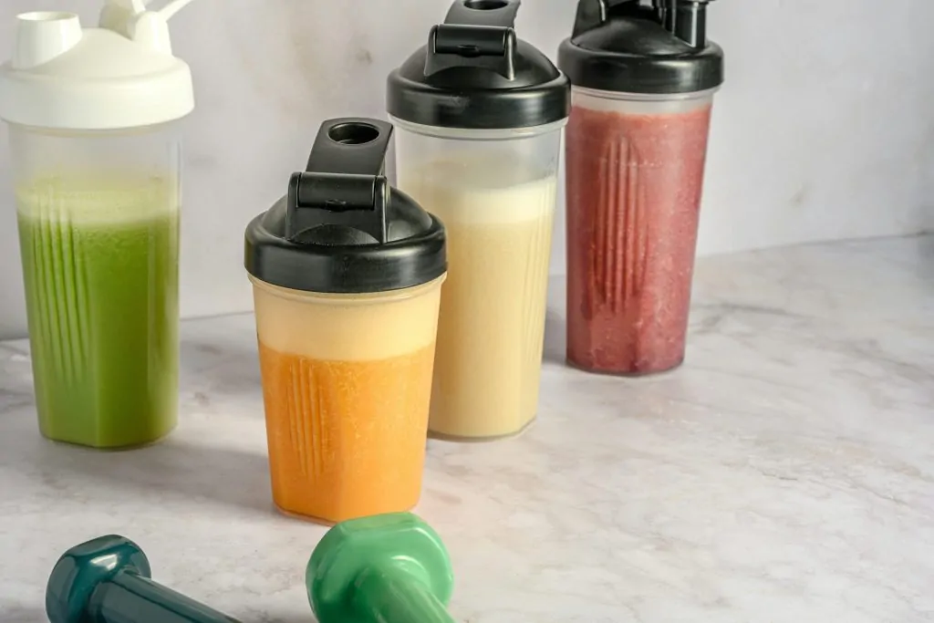 Vier drinkbekers met verschillende kleuren mixdranken en twee dumbbells op een marmeren oppervlak.
